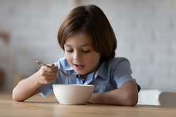 Little hungry cute boy sit at dinner table in kitchen holds spoon eating porridge or whole grain dry cereal with pleasure and appetite, enjoy balanced food. Nutrition, tasty healthy breakfast concept
