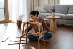 Full length smiling millennial generation indian woman sitting on warm heated floor, constructing wooden furniture in modern living room, fixing broken chair or table, improving interior at home.