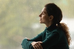 Music of rain. Side shot of serene latin woman relax by window with closed eyes listen sound of raindrops feel pleasure. Calm young lady enjoy breathing fresh ozonized air after rainstorm. Copy space.