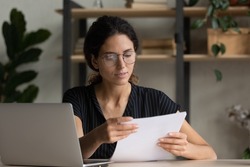 Important papers. Serious hispanic female manager in glasses engaged in office paperwork study project documentation cover letter. Young woman hr reading resume thinking on applicant cv at workplace