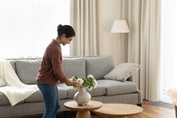 Smiling young indian ethnicity woman homeowner putting beautiful flowers in vase on coffee table in modern living room, enjoying decorating own apartment, improving styling interior on weekend.