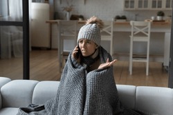 Unhappy young woman wrapped in blanket making phone call, freezing, sitting on couch, dissatisfied young female renter tenant complaining of low temperature in apartment, cold at home concept