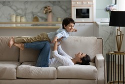 Happy young Indian mother lifting in air on straight arms joyful cute small child son, lying on cozy couch. Joyful family practicing balance doing yoga exercises, having fun playing at home.