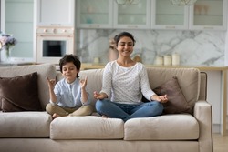 Happy small child boy sitting in lotus position on comfortable couch with joyful Indian mother, practicing together yoga breathing exercise, doing asanas, enjoying domestic healthcare activity.