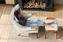 Top view peaceful African woman relax on armchair in modern cottage house with fireplace, put feet on comfy footstool enjoy comfort, rests in warm living room. No stress, air-conditioned home concept