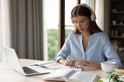 Focused engaged student girl in big headphones studying foreign language, listening audio lesson at laptop, reading notes out loud, doing exercises from open book, school, college homework task