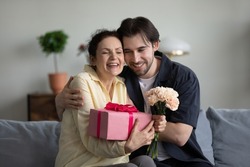 Joyful bonding middle aged senior woman feeling excited getting wrapped gift box and flowers from young smiling grownup son, sitting together on cozy couch, birthday or special occasion celebration.