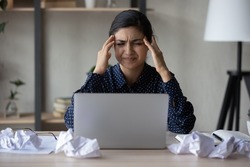 Unhappy annoyed Indian business woman feeling strong headache, anger, stress, nervous tension. Employee, office worker sitting at laptop and thrown papers, suffering from painful migraine