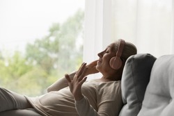 Side view happy old mature retired woman listening popular music in headphones, relaxing alone on cozy sofa, enjoying peaceful carefree weekend time alone at home, stress free leisure pastime.