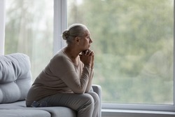 Pensive old mature woman looking in distance out of window, sitting on couch alone at home, suffering from negative thoughts or psychological problems, retirement lifestyle, ageing process concept.