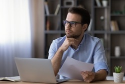 Thoughtful serious businessman in glasses touching chin, looking in distance, planning, holding document, sitting at desk with laptop, pensive man thinking, accountant working on financial report