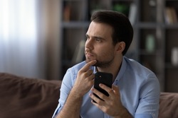 Head shot thoughtful serious man looking in distance, holding smartphone, touching chin, sitting on couch at home, confident businessman thinking pondering problem solving, making difficult decision