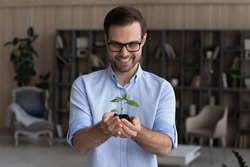 Smiling businessman wearing glasses holding small green plant sprout with soil, standing, happy entrepreneur employee with growing tree, startup project, profit, investment and growth concept