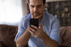 Close up thoughtful man looking at smartphone screen, touching chin, sitting on couch, pensive businessman thinking, reading news in email or message, hesitating and doubting, pondering offer