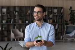 Dreamy smiling man in glasses holding green plant with soil, growing small tree, environment concept, happy pensive businessman entrepreneur with sprout in hands, investment and growth