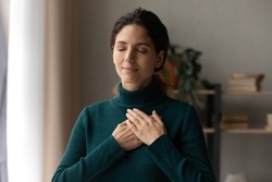 Head shot peaceful young attractive woman holding folded hands on chest, praying waiting for miracle, feeling thankful indoors. Sincere happy female volunteer showing kindness or expressing gratitude.