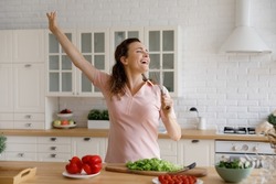 Overjoyed young beautiful woman singing favorite song in whisk as microphone, having fun cooking healthy vegetarian food alone in modern kitchen, enjoying domestic culinary hobby activity on weekend.