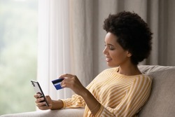 Happy young African American woman holding cellphone and bank credit card in hands, transferring money online, shopping goods in internet store, purchasing services, satisfied with secure payments.