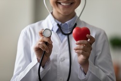 Close up cropped smiling young Indian female doctor in white medical coat holding stethoscope and small heart figure, recommending checkup for disease prevention or advertising cardiology services.