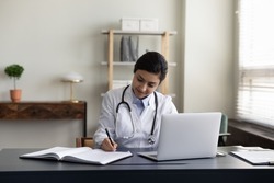 Happy millennial Indian female medical worker doctor in white uniform making notes in registration journal, managing appointments or prescribing illness treatment for patient, working on laptop.