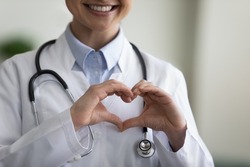 Close up happy young Indian female GP doctor therapist physician making heart gesture with fingers, showing love care support to patients, professional medical help, charity donation concept.