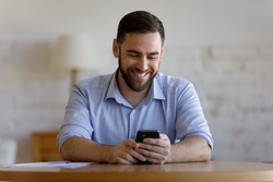 Happy cheerful millennial business man chatting online to client on smartphone at office workplace table, reading text message with good news, browsing internet, typing on mobile phone, smiling