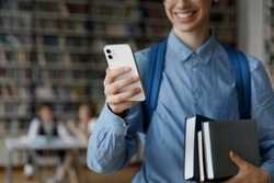Happy young gen Z student guy using online app, internet service on smartphone, holding stacked books, textbooks, smiling, visiting college library. Hand holding mobile phone close up