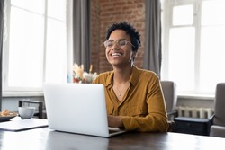 Cheerful short haired African employee sitting at workplace with laptop in office, typing, looking at camera, smiling, laughing. Millennial worker, working business woman in casual head shot portrait
