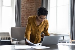 Busy millennial African entrepreneur woman reviewing online reports, using laptop computer. Freelance employee, businesswoman working in home office, doing paperwork job, standing at table