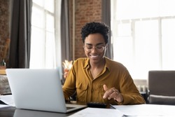 Happy young Afro American entrepreneur woman in glasses counting profit, on calculator at laptop computer, analyzing benefits, enjoying financial success, job high result, smiling