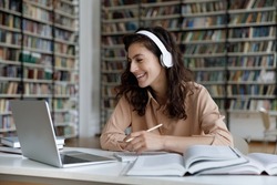 Happy engaged gen Z student girl watching webinar, attending online class, learning conference, talking to teacher on video call, laughing, writing, studying in library with headphones, laptop, books