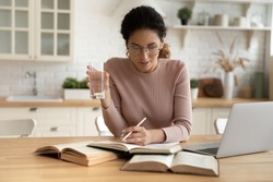 Serious young Latina woman glasses holds glass of water while studying use textbooks read educational books seated at dining table, learns prepare for university exam. Self-education at home concept