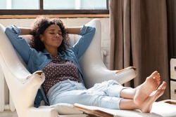 Full length millennial African American woman homeowner daydreaming in comfortable armchair with legs on footstool, breathing fresh air, sleeping resting, enjoying peaceful leisure time at home.