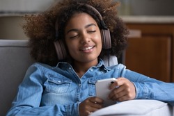 Smiling peaceful young gen Z teen Black girl wearing big wireless headphones, using smartphone, enjoying relaxing romantic music resting on comfortable couch at home with closed eyes