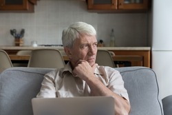 Pensive stressed mature 70s man holding laptop computer, looking away in deep sad thoughts, thinking over bad concerning news, health problems, sitting on home couch, feeling worried, frustrated