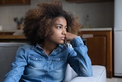 Frustrated pensive teenage African girl sitting on couch at home, looking away, thinking over heartbreak problems, offence, feeling bored, suffering from depression. Youth problems concept