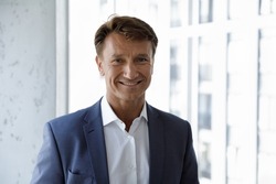 Happy mature male business leader head shot portrait. Confident middle aged 50s businessman, CEO, executive in formal suit looking at camera, smiling, standing at office window. Job success concept