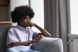 Thoughtful sad teen Black guy with retro Afro hairstyle sitting on sofa at home, looking away, thinking, feeling anxious, bored, concerned, suffering from depression. Youth problems concept