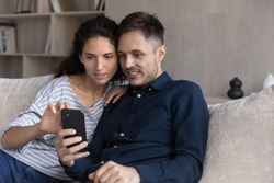 Focused millennial couple using online service on smartphone together, sharing mobile phone shopping on internet application. Young husband and wife relaxing on couch at home with digital gadget