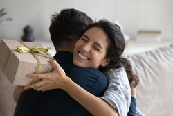 Grateful happy young woman holding festive wrap, hugging beloved husband, boyfriend, thanking for present. Sweet millennial couple celebrating anniversary, 8 march, birthday, exchanging gifts