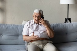 Worried concerned senior elder man talking on cellphone to doctor, feeling unwell, calling for emergency, ambulance, sitting on sofa at home, contact helpline, using mobile phone