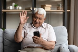 Happy friendly senior 80s grandpa man speaking on video conference call on smartphone, waving greeting hand hello from home couch, talking to grandchildren online. Distant communication concept
