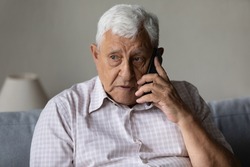 Serious concerned old grandpa talking on mobile phone, calling for emergency, ambulance. Senior 80s man speaking on cellphone, sitting on couch, at home, contact family on distance