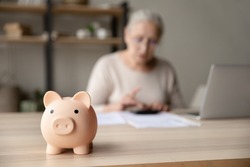 Toy pink piggy bank on work table of senior tenant, homeowner woman. Elderly lady using calculator, counting savings, taxes. Finance management, financial insurance, personal budget concept