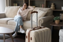 Focus on beige suitcase with blurred carefree young woman sitting on sofa on background. Joyful refreshed millennial Hispanic lady satisfied with vacation travel, relaxing alone in modern hotel room.