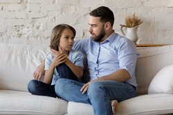 Serious father listen to his pre-teen little son talking seated on sofa at home, speaking spend time together at home. Cute boy share problems, ask advice to dad. Communication, care and trust concept