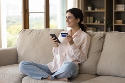 Dreamy happy young woman feeling excited purchasing favorite goods or service online with discount in mobile shopping application, satisfied with secure money transfer, sitting on couch at home.
