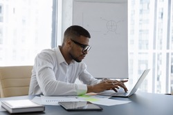 Concentrated young African American businessman employee manager in eyeglasses looking at laptop screen, typing on keyboard working on online project, web surfing information or analyzing statistics.