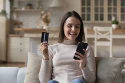 Happy young woman holds smartphone and credit card make purchase remotely, buy goods on internet, transfer money successfully use electronic services. E-shopping, secure pay through e-bank app concept