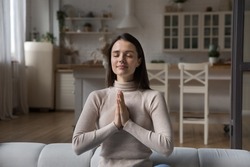 Young woman sit on sofa with eyes closed and palms folded in namaste or prayer gesture, asking sincerely for miracle and luck pleading for innermost, express gratitude. Faith in God, religion concept